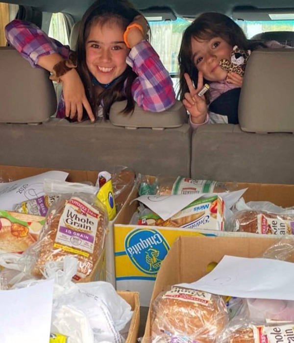 Photo from the trunk of an SUV-type car. Trunk is full of boxes of nutritious groceries. Two White girls ages 10-12 are smiling from the car's middle seats and throwing peace signs with their fingers.