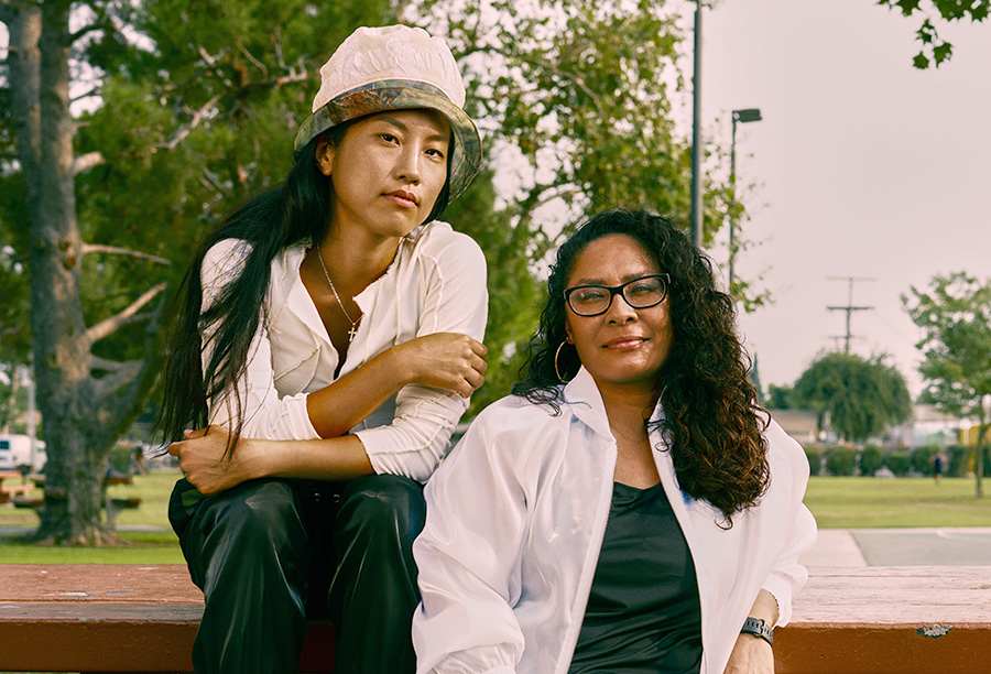 Photo of two female presenting people, one Asian and one Latina, sitting on a picnic table in a park. Their facial expressions are mildly defiant yet positive, showing strength and resilience.
