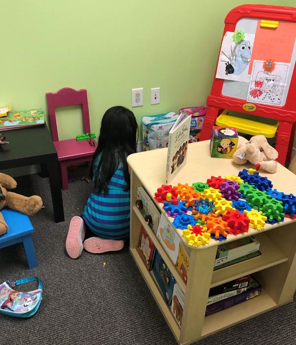 Photo of back of a young girl with long dark hair playing in a playroom surrounded by colorful toys.
