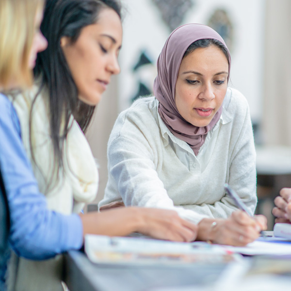 Photo of 3 women collaborating at a table. The woman at the focal point of the image is wearing a head scarf and writing on a piece of paper at the center of the 3 women.
