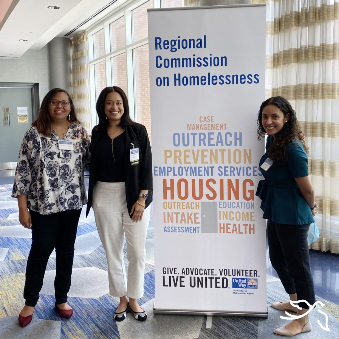 Three women of color smiling and standing in front of a tall freestanding banner titled "Regional Commission on Homelessness"