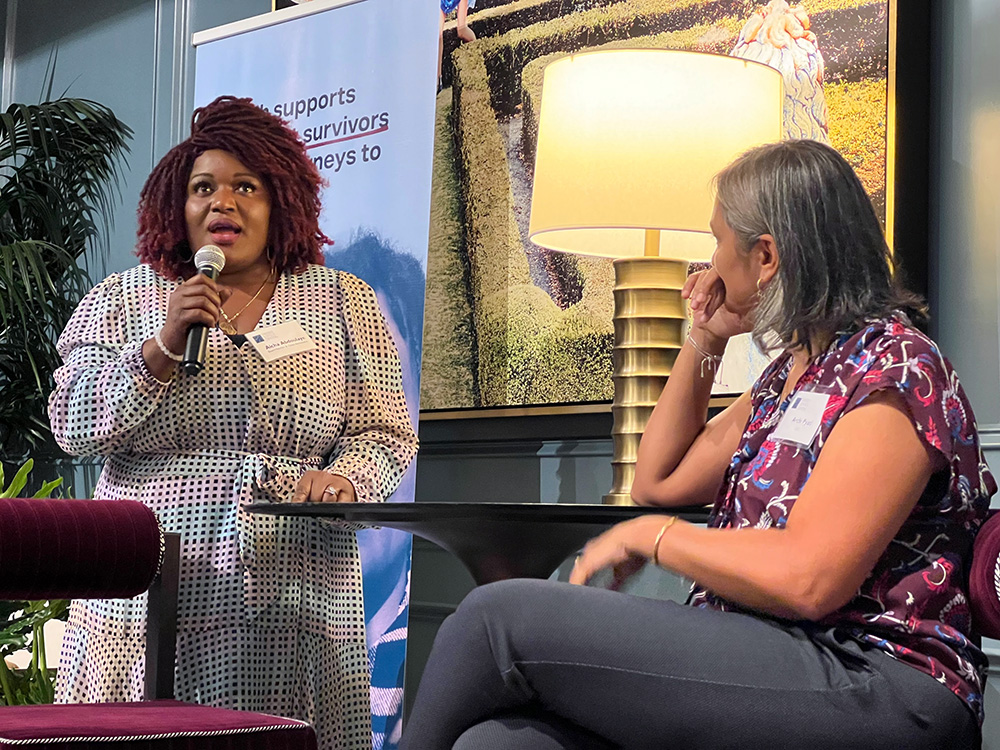 Woman of color talking with microphone. Caucasian woman sitting next to her and looking with attention.