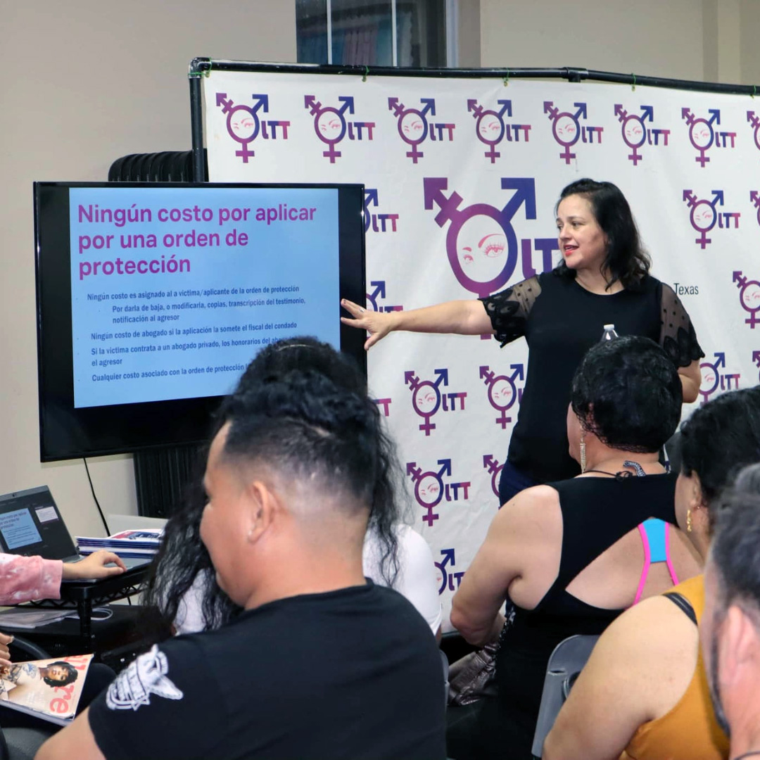 Woman giving a presentation, gesturing with her arm toward a screen with a slide titled "Ningún costo por aplicar por una orden de protección" (No cost to apply for a protection order)