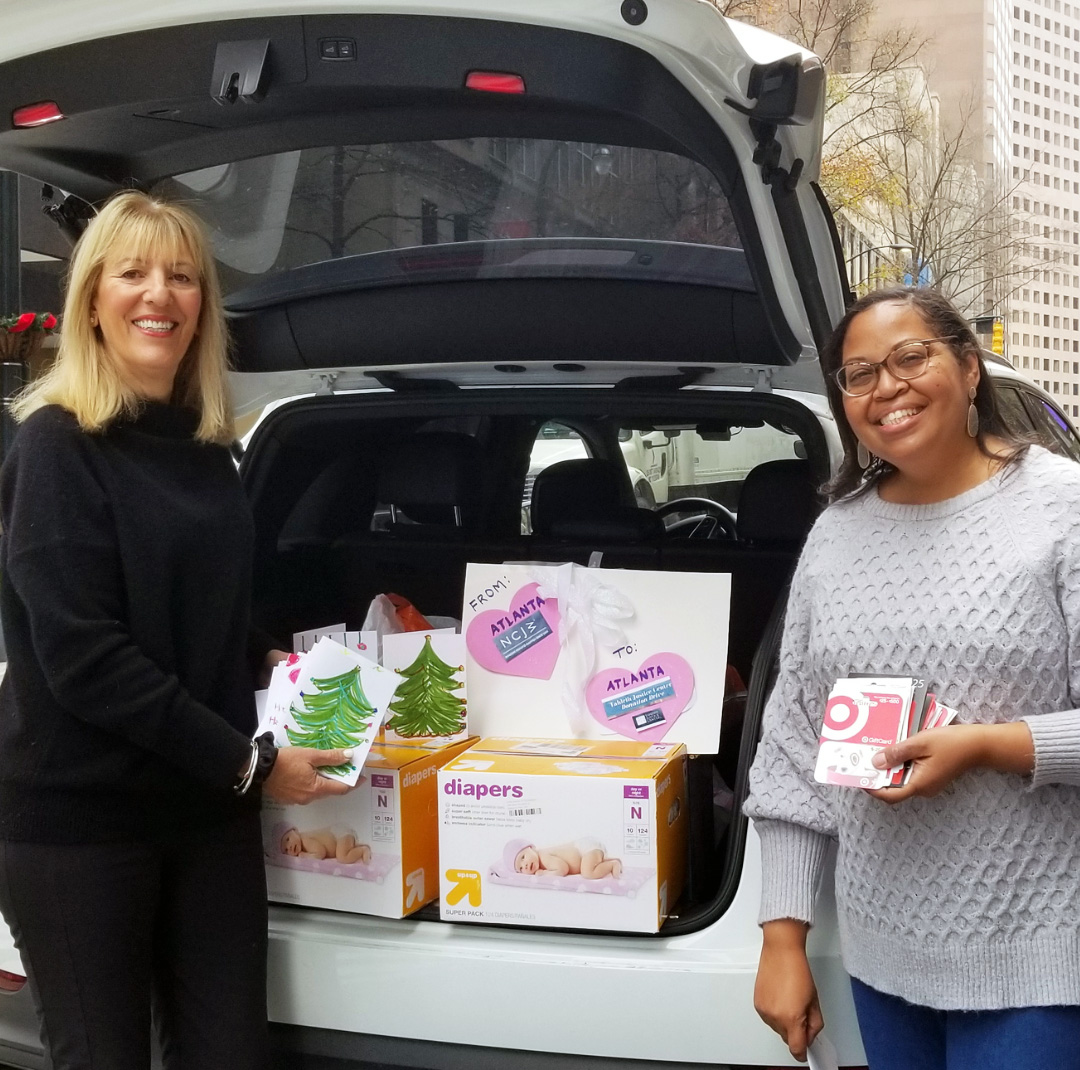 Two women smiling, standing in front of an open trunk of an SUV filled with boxes of diapers and gifts to donate.