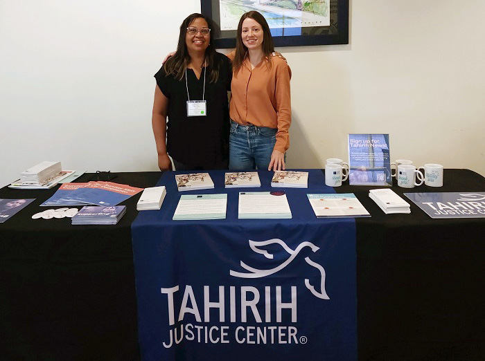 BIPOC woman and caucasian woman standing in front of a table with printed materials and a Tahirih Justice Center tablecloth banner.