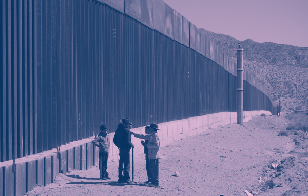 Immigrants stand next to a border fence at the U.S. Mexico border.