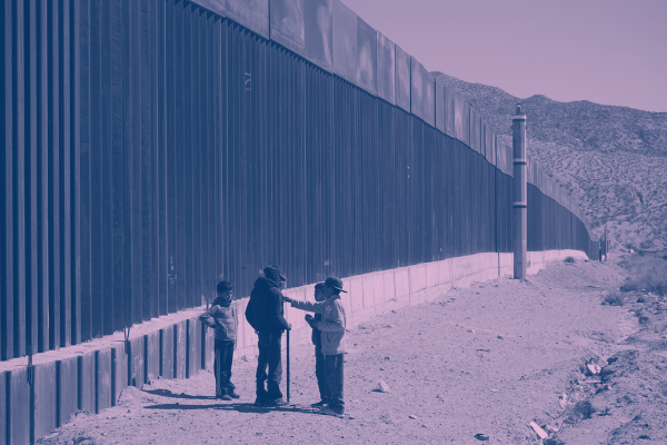 Immigrants stand next to a border fence at the U.S. Mexico border.