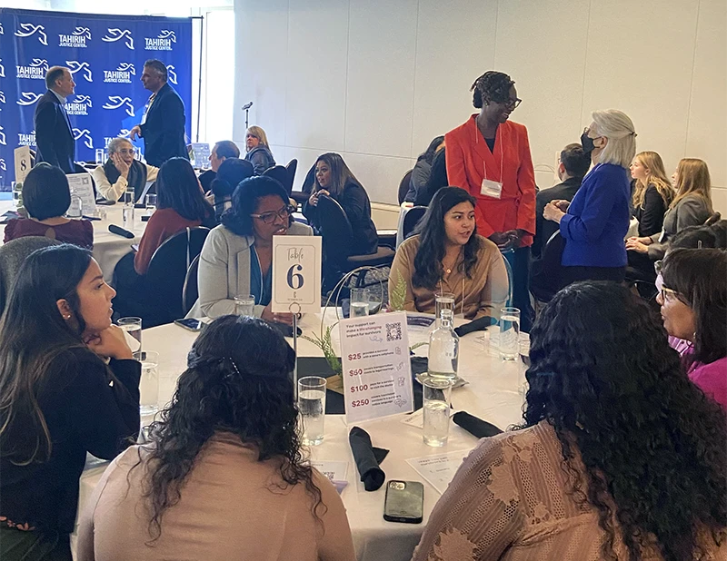 Luncheon setting with Tahirih banner in the background, a group of women sitting around a table in the foreground engaged in conversation