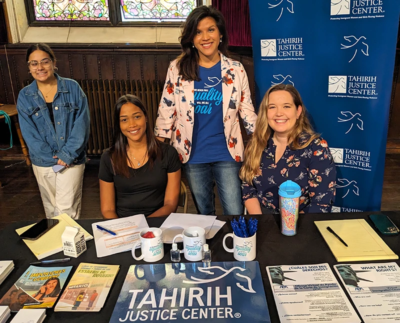Four women in front of a Tahirih Justice Center banner, sitting behind a table containing Tahirih materials in English and Spanish
