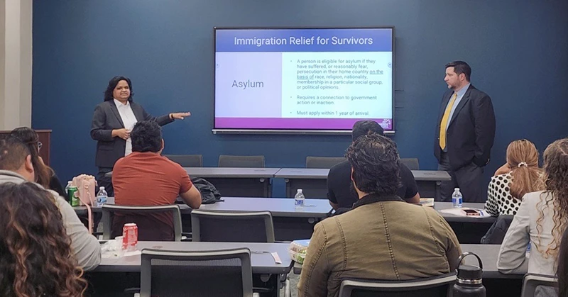 Woman and man stand at the front of the room with presentation of asylum topics. Mixed audience listens.