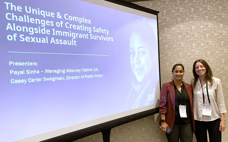 Two women standing in front of a presentation screen with title "The Unique and Complex Challenges of Creating Safety Alongside Immigrant Survivors of Sexual Assault"