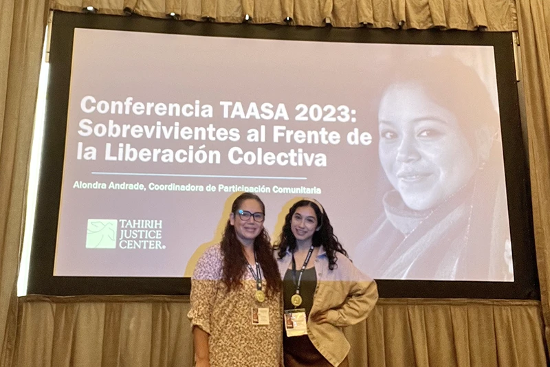 Two women in front of a presentation screen titled "Conferencia TAASA 2023: Sobrevivientes al Frente de la Liberación Colectiva"