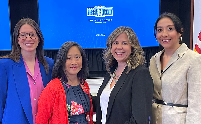 Four women stand in front of presentation screen with The White House displayed and American flag to the side.
