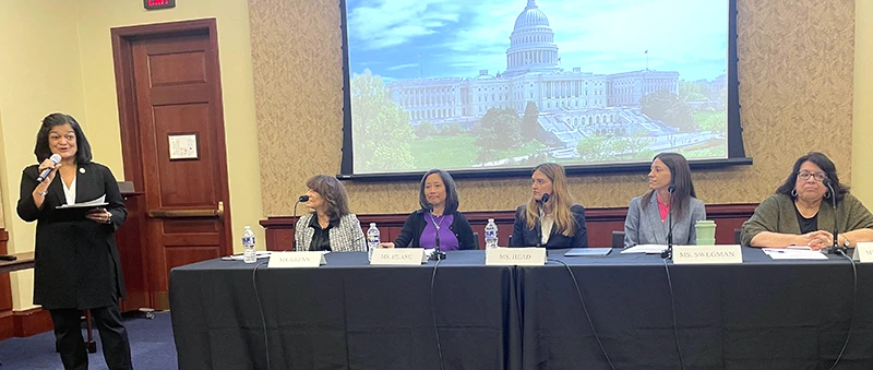 Five women on a panel in front of a photo of the U.S. Capitol building listen to another woman speak with a microphone.