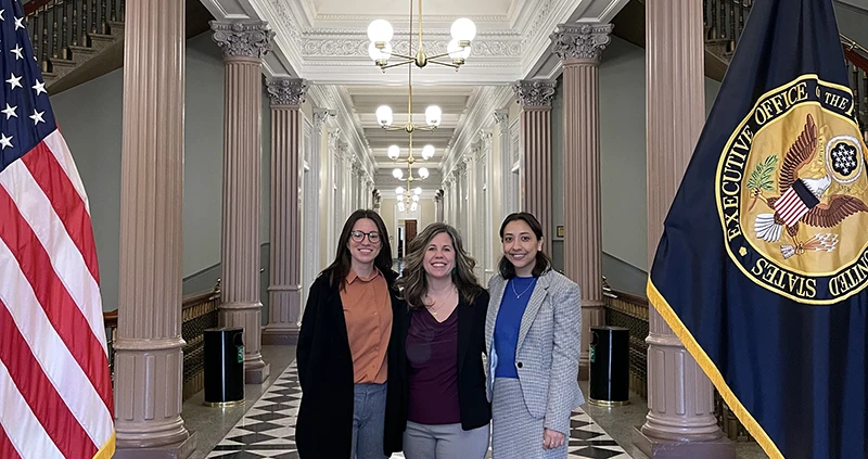 Three women stand in large hallway with stone pillars and American flag and Executive Office flag on either side.