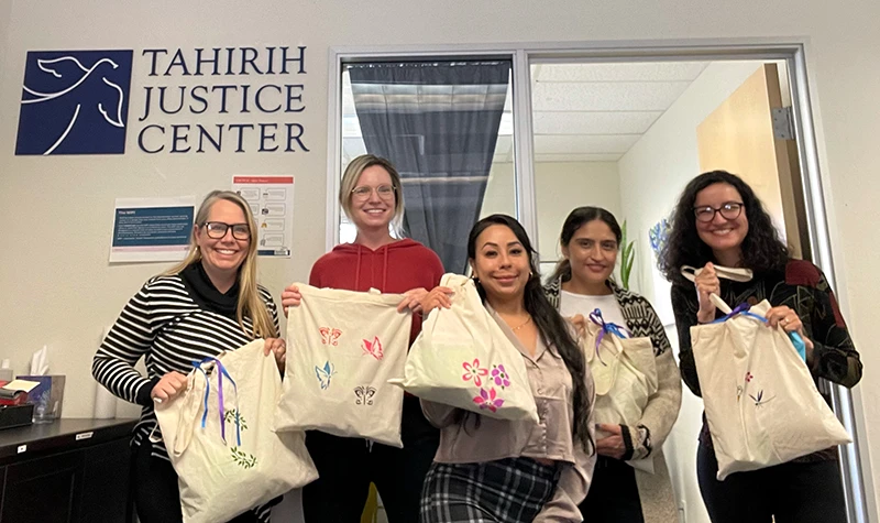 Five women in office with Tahirih Justice Center logo on the wall holding up bags and smiling
