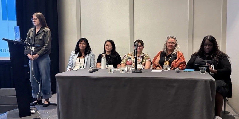 Women on a stage for a panel discussion. One woman is speaking at a podium.