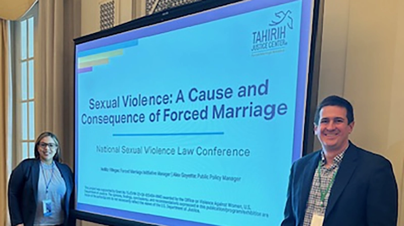 Man and woman stand smiling in front of a large presentation screen with title "Sexual Violence: A Cause and Consequence of Forced Marriage"