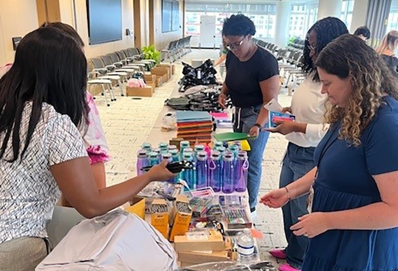 Women standing along a long table selecting backpacks, school supplies, water bottles and other products to assemble backpacks for children.